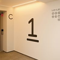 Černá podtržená číslice 1 na stěně, označení patra, plotrované navigační piktogramy, dveře výtahu s čísly pater, písmeno C na stěně