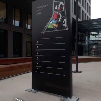 Černý plechový totem na betonové ploše před černou administrační budovou, orientační plán PALMOVKA OPEN PARK, čísla budov