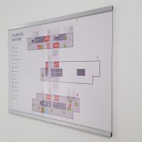 Orientační systém Floor 6 NATURE na Dibond desce s plexisklem, nalepeno na bílé zdi, vpravo detail černých písmen, číslovek a šipek nalepených na zdi