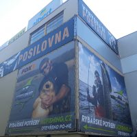 Roh budovy, vpravo i vlevo pokryt bannerem s reklamou na rybářské potřeby, vpravo šedá část budovy, nahoře další část budovy a jiné reklamní bannery