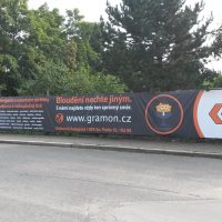 Exteriér, v popředí asfaltová silnice, v pozadí chodník, za ním na plotě banner, v pozadí stromy