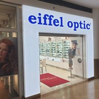 Podsvícená bílá brána do prodejny nahoře s modrým nápisem eiffel optic, v prostředí nákupního centra, uvnitř prodejny vitríny s produkty, na zemi červený koberec