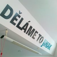 Slogan DĚLÁME TO JINAK vyřezaný z černé a modré plotrové folie nalepený na bílé stěně u stropu, svítidlo