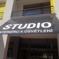 Sluneční clona polepená černou samolepicí folií s bílým nápisem STUDIO INTERIÉRU A OSVĚTLENÍ, detail bílé budovy s balkonem