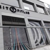 Černé 3D logo FITLAB na šedé stěně budovy, okna polepená barevnou děrovanou folií