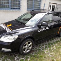 Zboku vyfocené černé osobní auto, na kapotě bílý nápis CONSTUCTING a oranžové logo nad ním, na dveřích u řidiče bílý nápis CONSTRUCTING.CZ, na blatníku nad zadním kolem oranžový polep, v exteriéru, před budovou