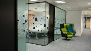Skleněné příčky tvořící zasedací místnost v v kancelářské budově, polep pískovanou folií s modrými kruhy, šedý koberec, černo zelené křeslo