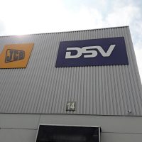 Dvě reklamní desky s logy JCB a DSV, instalované na kovové budově, pohled zespoda, v pozadí obloha s mraky