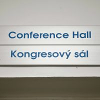 Dva informační kovové štítky nad sebou, modré nápisy Conference Hall a Kongresový sál, přišroubované na bílé zdi, pohled zespodu