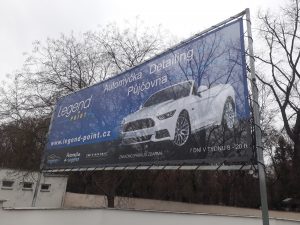 Exteriér, na kovové konstrukci napnutý modrý banner s motivem bílého auta, bílé nápisy, v pozadí opadané stromy