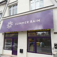 Velká dibond deska polepená fialovou folií s bílým logem SUMMER RAIN instalovaná nad výlohou provozovny, bílá budova s okny