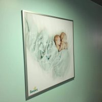 Světlá modro-zelená stěna, uprostřed stříbrný rám s reklamou na Pampers, vpravo v pozadí dveře