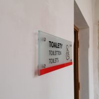 Informační panel z pískovaného konexového skla, černý nápis TOALETY, TOILETTEN a TOILETS s piktogramem handicapované osoby, připevněno distančními šrouby na zdi vedle dveří
