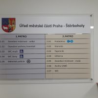 Skleněný informační panel Úřadu městské části Praha-Štěrboholy, znak městské části, značneí úřadu a místností, přimontováno na bílé zdi, pohled zepředu