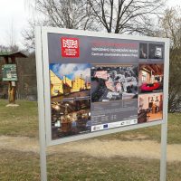 Hliníkový stojan s cedulí, pozvánka Centra stavitelského dědictví Plasy, fotky expozice, v pozadí přírodní informační totem