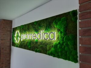 Tabule obdélníkového tvaru se zelenými výplněmi mechu, uprostřed s šedým nápisem promedica a šedým okrouhlým logem, nápis i logo bíle podsvíceno, na bílé stěně, vpravo i vlevo cihlové sloupy