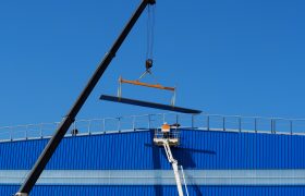 Rameno jeřábu zdvihá traverzu na střechu modré budovy, technik v reflexní vestě na plošině, modrá obloha