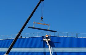 Rameno jeřábu zdvihá traverzu na střechu modré budovy, technik v reflexní vestě na plošině, modrá obloha