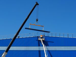 Rameno jeřábu zdvihá traverzu na střechu modré budovy, technik v reflexní vestě na plošině, modrá obloha