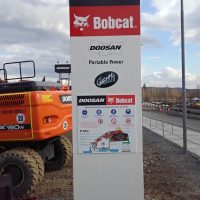 Vysoký totem s logem DOOSAN a Bobcat, cedule s orientačním plánem, zadní část oranžového stavebního stroje na štěrkové ploše