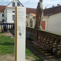 Bílý totem se zlatým pruhem, černým textem a orientačním plánem v areálu kláštera, socha světce na kamenném zábradlí, lavičky