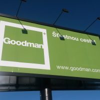 Pohled zespodu na dvojitý reklamní billboard firmy Goodman, zelenobíle potištěná dibond deska, černý rám a sloup, troje světla instalovaná na vrchu